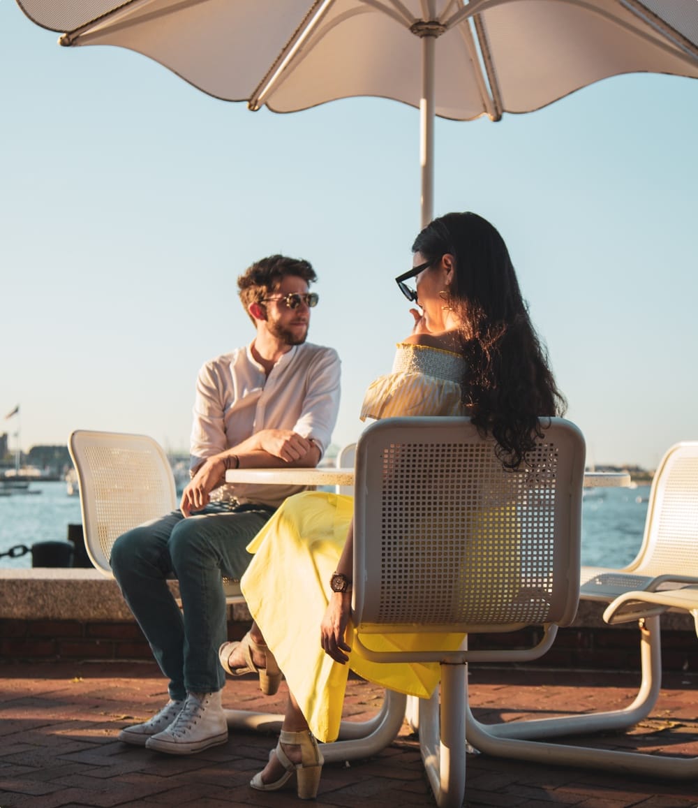 A couple sits together at a table with an umbrella by the water near Ora Seaport Apartments Boston