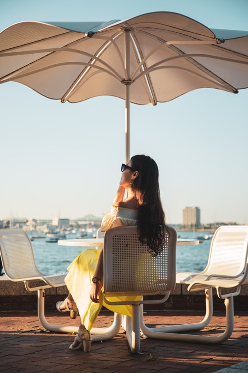 A woman in sunglasses sits at a table by the water near Ora Seaport luxury apartments in Boston